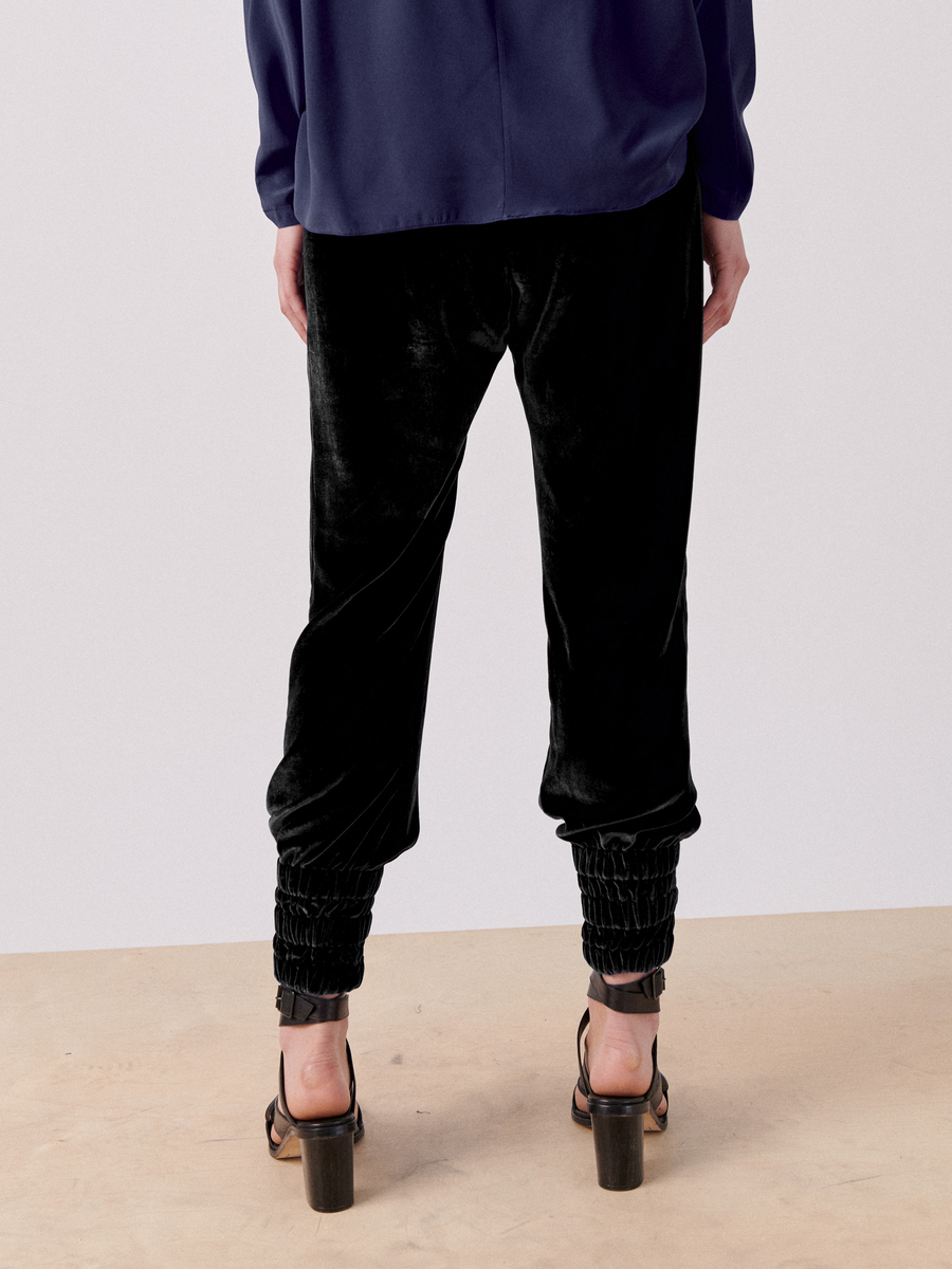 A person stands facing away, wearing the Zero + Maria Cornejo Tri Tabi Pant with a banded elastic waist and gathered cuffs, paired with high-heeled sandals. The individual is also wearing a dark blue top with long sleeves. The background is a minimalist indoor setting with a light floor and plain walls.