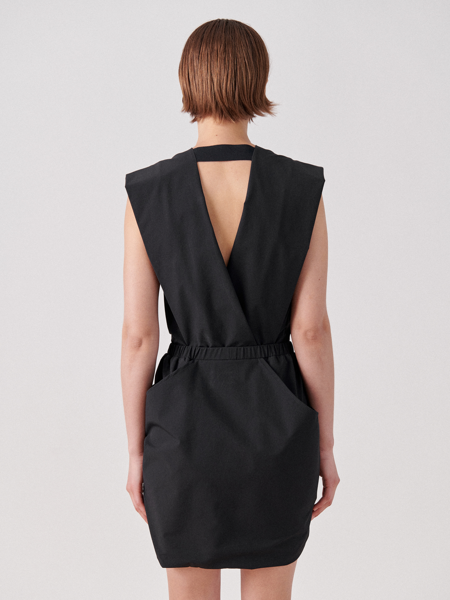 A person with short hair is wearing a sleeveless black Mini Mackie Dress by Zero + Maria Cornejo, shown from the back. The dress features a deep V-cut and an elastic waistband. The person's arms hang naturally by their sides, and the background is light gray.