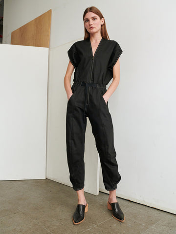 A woman stands against a minimalistic indoor background, wearing the sleek black Strap Akeo Jumpsuit by Zero + Maria Cornejo, made from GOTS-certified cotton. The jumpsuit features short sleeves and ankle cuffs. She pairs the outfit with black pointed-toe mules. Her long brown hair is parted down the middle, and she has her hands in the jumpsuit's pockets.