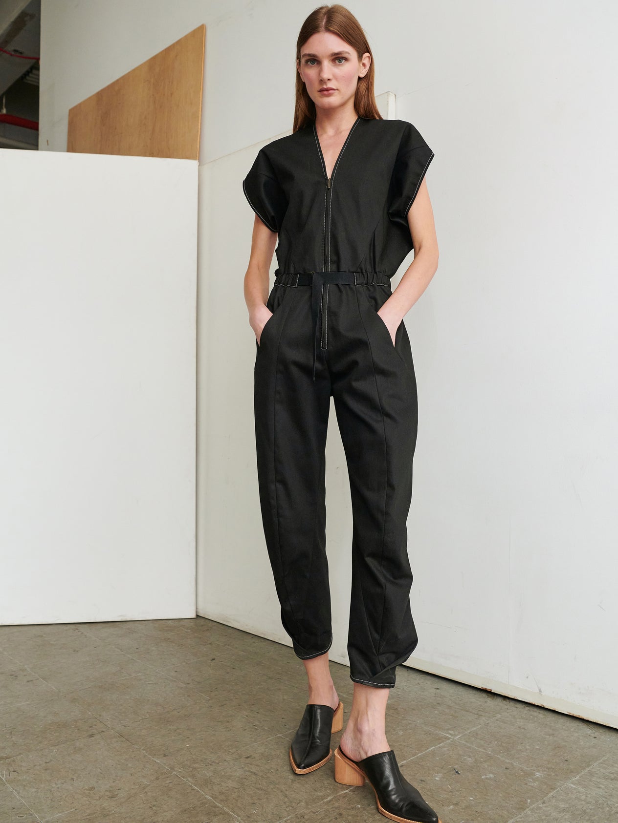 A person with long brown hair stands against a plain white wall, wearing the dark Strap Akeo Jumpsuit made of 100% organic cotton denim from Zero + Maria Cornejo. The jumpsuit features short sleeves, a zippered front, and is belted at the waist. They have their hands in their pockets and are wearing black slip-on shoes.
