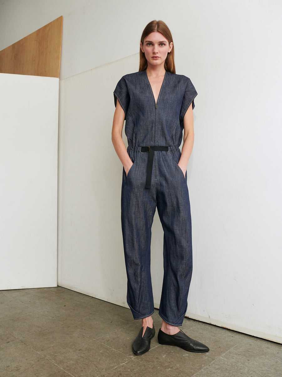 A person with long brown hair stands against a plain white wall, wearing the dark Strap Akeo Jumpsuit made of 100% organic cotton denim from Zero + Maria Cornejo. The jumpsuit features short sleeves, a zippered front, and is belted at the waist. They have their hands in their pockets and are wearing black slip-on shoes.