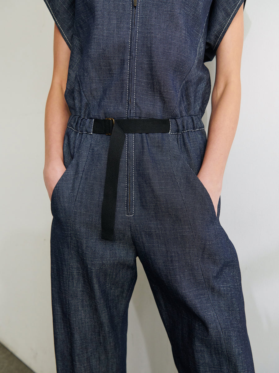 A person wearing a Strap Akeo Jumpsuit by Zero + Maria Cornejo, crafted from GOTS-certified cotton. The dark, organic denim jumpsuit features short sleeves, a front zipper, and a black belt at the waist. With their hands in the pockets and the photo cropped just above the shoulders.