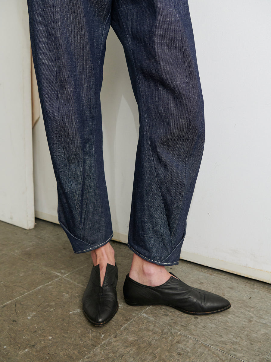 A person wearing a dark blue Strap Akeo Jumpsuit from Zero + Maria Cornejo, crafted from GOTS-certified cotton and featuring unique leg stitching, stands on a textured gray floor near a white wall. They are also wearing black pointed-toe slip-on shoes.