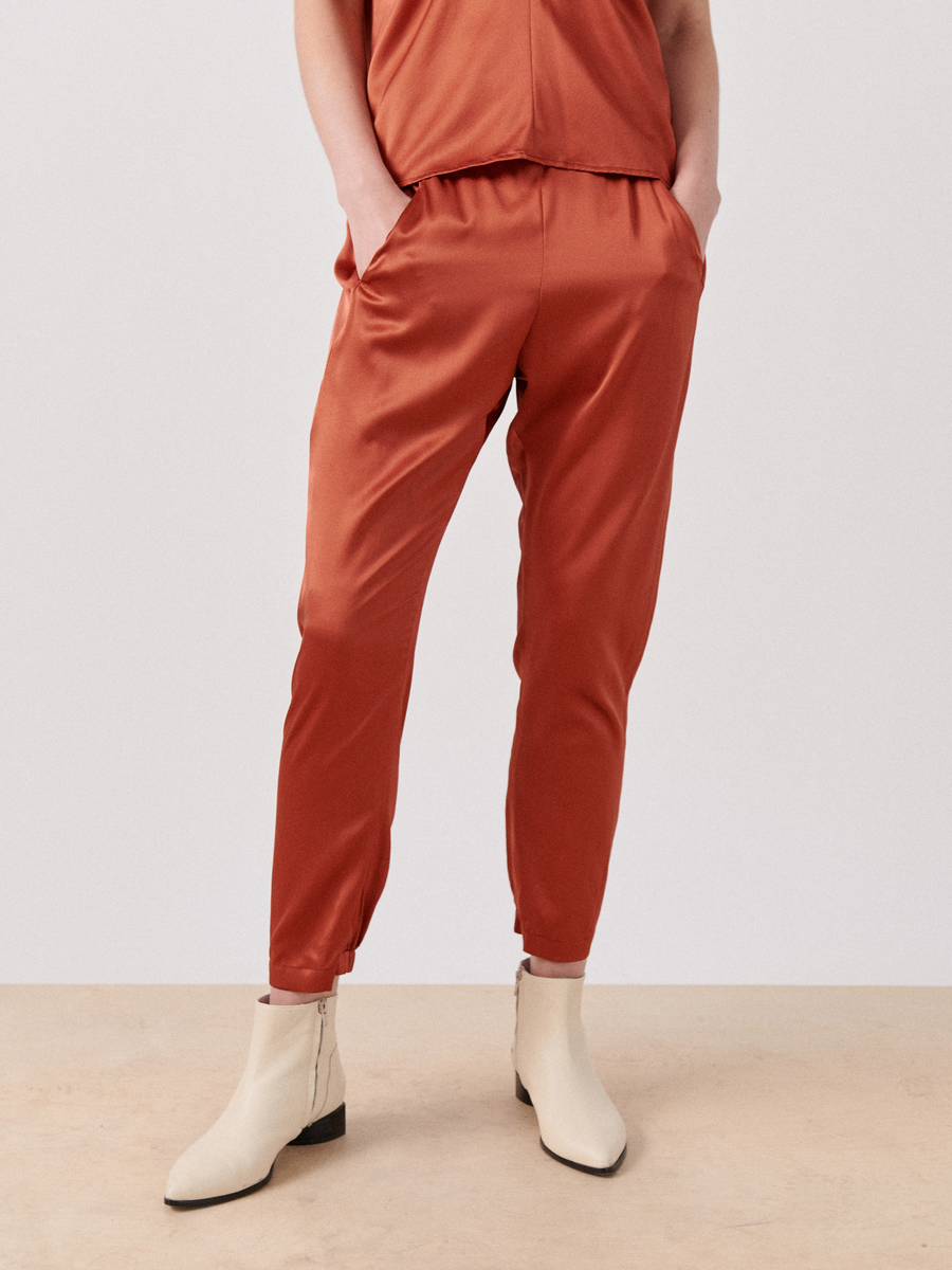 Person standing in relaxed orange satin Gabi Trouser by Zero + Maria Cornejo with an elastic waist and hands in pockets, paired with white ankle boots and a matching orange top against a plain light background.