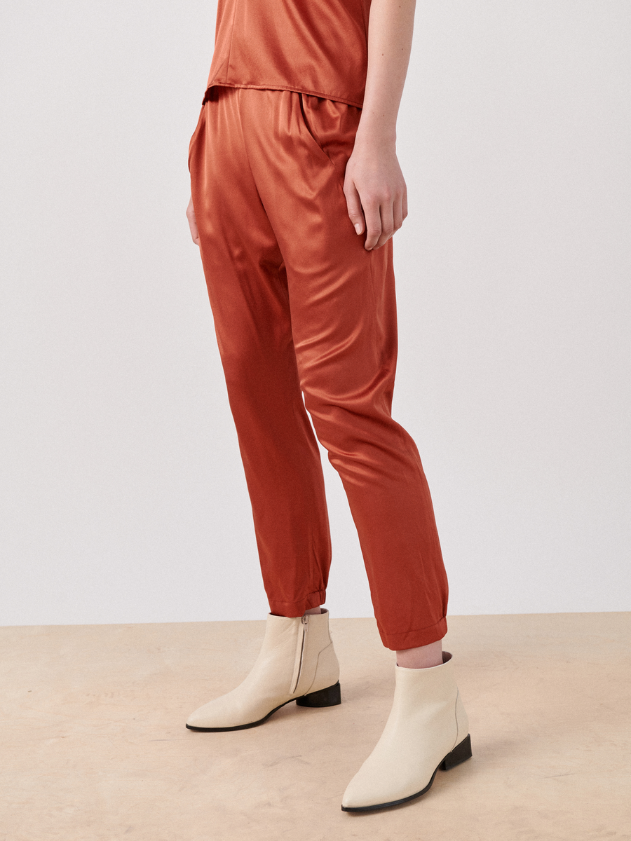 A person stands on a beige floor, wearing rust-colored Gabi Trouser by Zero + Maria Cornejo and a matching top made of stretch silk charmeuse. Their feet are clad in off-white ankle boots with low heels. The background is plain and light-colored, creating a minimalist setting.