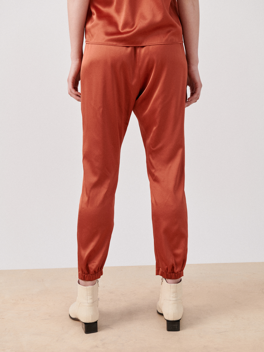 A person stands with their back to the camera, wearing rust-colored Gabi Trousers from Zero + Maria Cornejo with a relaxed fit and elastic waist elasticated cuffs. They are paired with cream-colored boots with black soles. The background is plain and neutral.