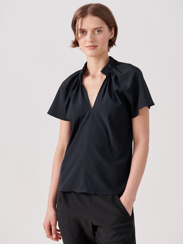 A person with short brown hair is wearing a black Ruched Stella Top by Zero + Maria Cornejo. They are standing against a plain white background with one hand in their pocket, looking towards the camera.