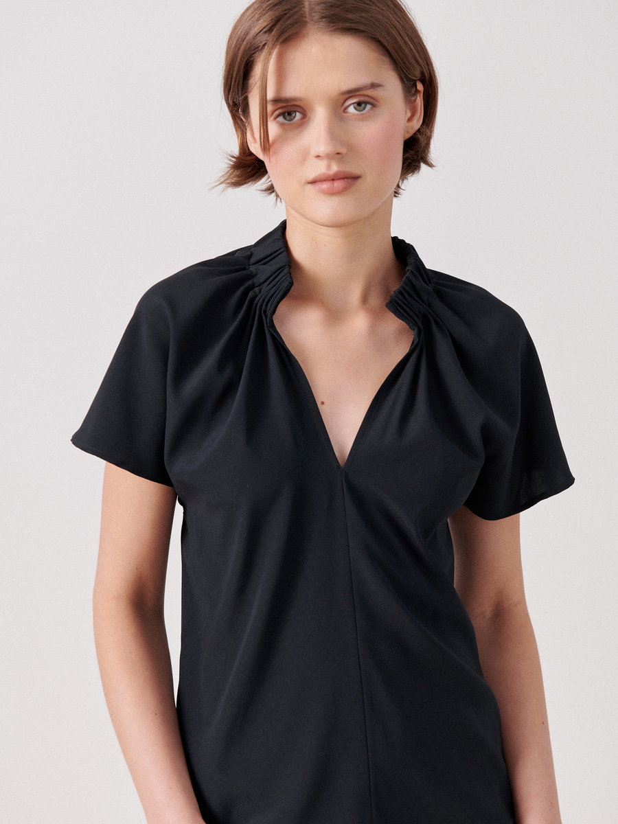 A person with short, light brown hair is wearing a black Ruched Stella Top by Zero + Maria Cornejo with gathered detailing at the collar and short sleeves. They are standing against a plain, light gray background, looking directly at the camera with a soft expression.