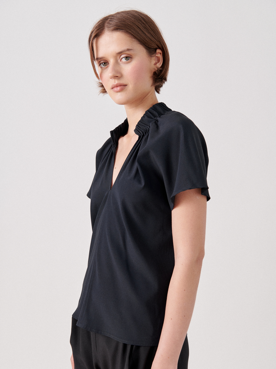A woman with short brown hair is wearing a black Ruched Stella Top by Zero + Maria Cornejo, crafted from stretch silk charmeuse with a deep V-neck and subtle gathered detailing at the shoulders. She is looking at the camera with a neutral expression against a plain light background.