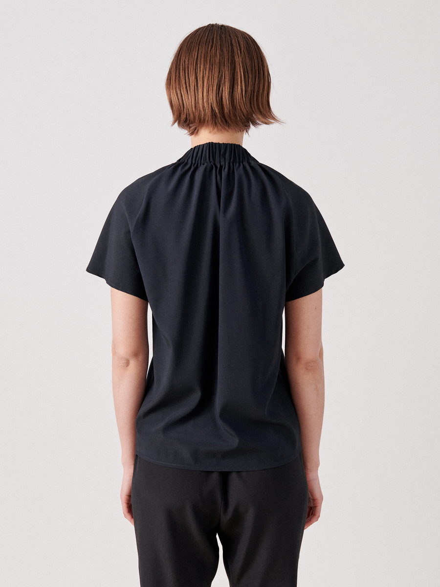 A person with short brown hair is shown from the back, wearing a black Ruched Stella Top by Zero + Maria Cornejo with black pants. The plain background is light gray.