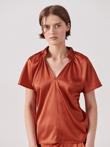 A woman wearing a rust-colored, short-sleeve, V-neck Ruched Stella Top by Zero + Maria Cornejo with gathered detailing near the neckline stands against a plain light gray background. She has short brown hair and a neutral expression.