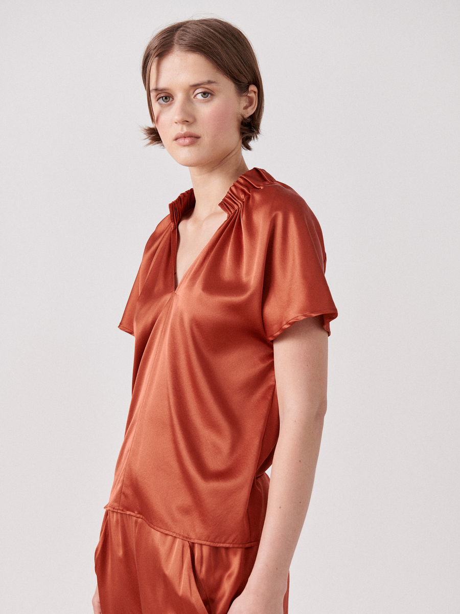 A person with short hair stands against a plain backdrop, dressed in a shiny, loose-fitting burnt-orange outfit. The ensemble includes the Zero + Maria Cornejo Ruched Stella Top with matching bottoms. They have a neutral expression, and the garment features subtle gathered details at the collar.