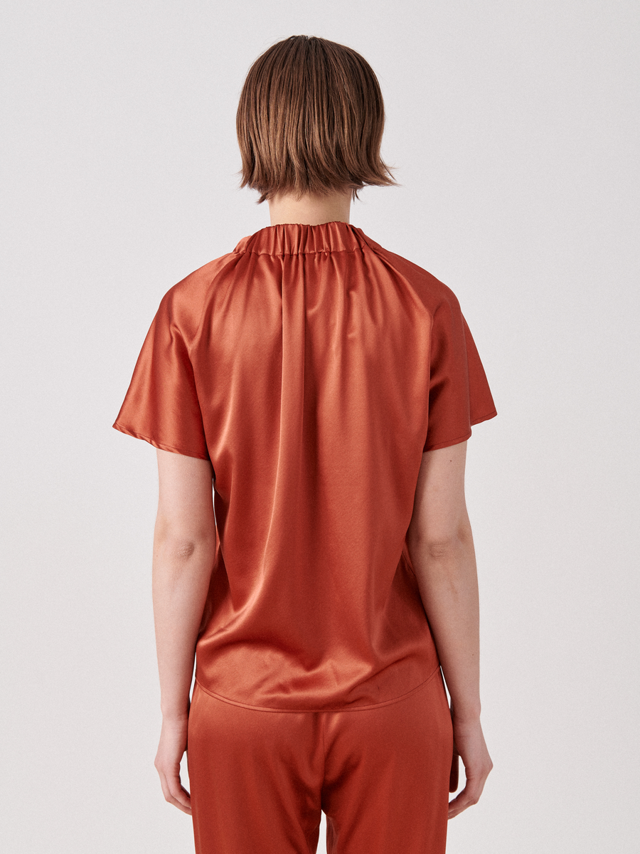 A person with short brown hair is facing away from the camera, wearing a short-sleeved, shiny rust-colored Ruched Stella Top by Zero + Maria Cornejo and matching trousers. The top has a gathered neckline. The background is plain and light-colored.
