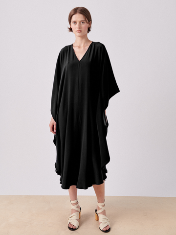 A person with short, dark hair stands against a plain background wearing the Circle Caftan from Zero + Maria Cornejo—a loose-fitting black silk charmeuse dress with a V-neck and draped sleeves—and beige, strappy sandals.