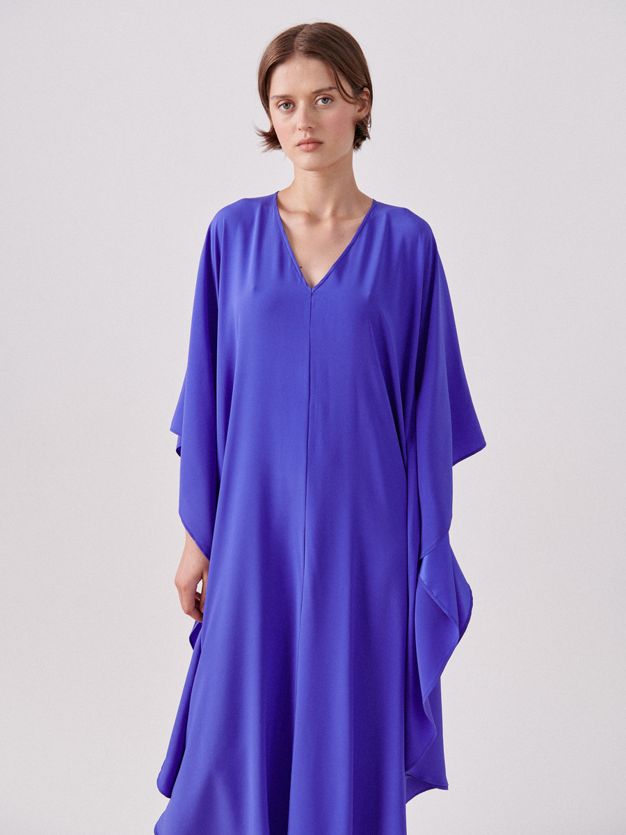 A person with short hair is wearing a shimmering, bright blue Circle Caftan by Zero + Maria Cornejo with a V-neckline and wide sleeves. They are standing against a plain, light-colored background and looking directly at the camera.