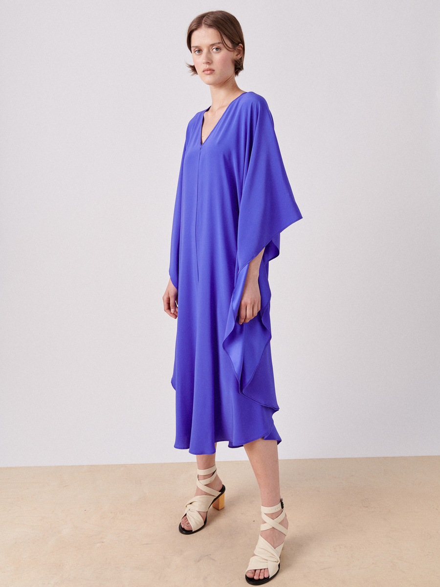 A person stands against a plain background, wearing a vibrant blue Circle Caftan with wide sleeves. They have short, light brown hair and are wearing cream and black strappy sandals. The Circle Caftan by Zero + Maria Cornejo, made from stretch silk charmeuse, reaches mid-calf and has a V-neckline with a shimmering appearance.