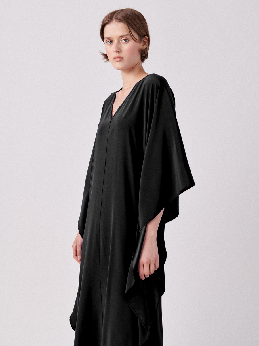 A person with short hair wears a flowing, black, V-neck Circle Caftan by Zero + Maria Cornejo. They stand against a plain white background, gazing forward with a neutral expression. The wide sleeves and draped silhouette create a relaxed fit, giving the garment a casual yet elegant look.