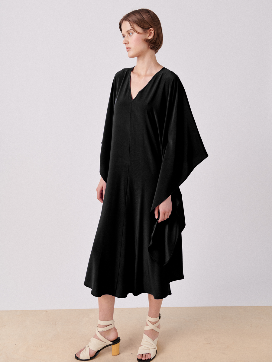 A person stands against a plain background wearing the Circle Caftan by Zero + Maria Cornejo, a flowy, mid-length black dress with a draped silhouette. The V-neckline and wide sleeves enhance its elegance. They are also wearing light-colored, open-toe heeled sandals with crisscross straps. The person has short, brown hair and looks to the side.