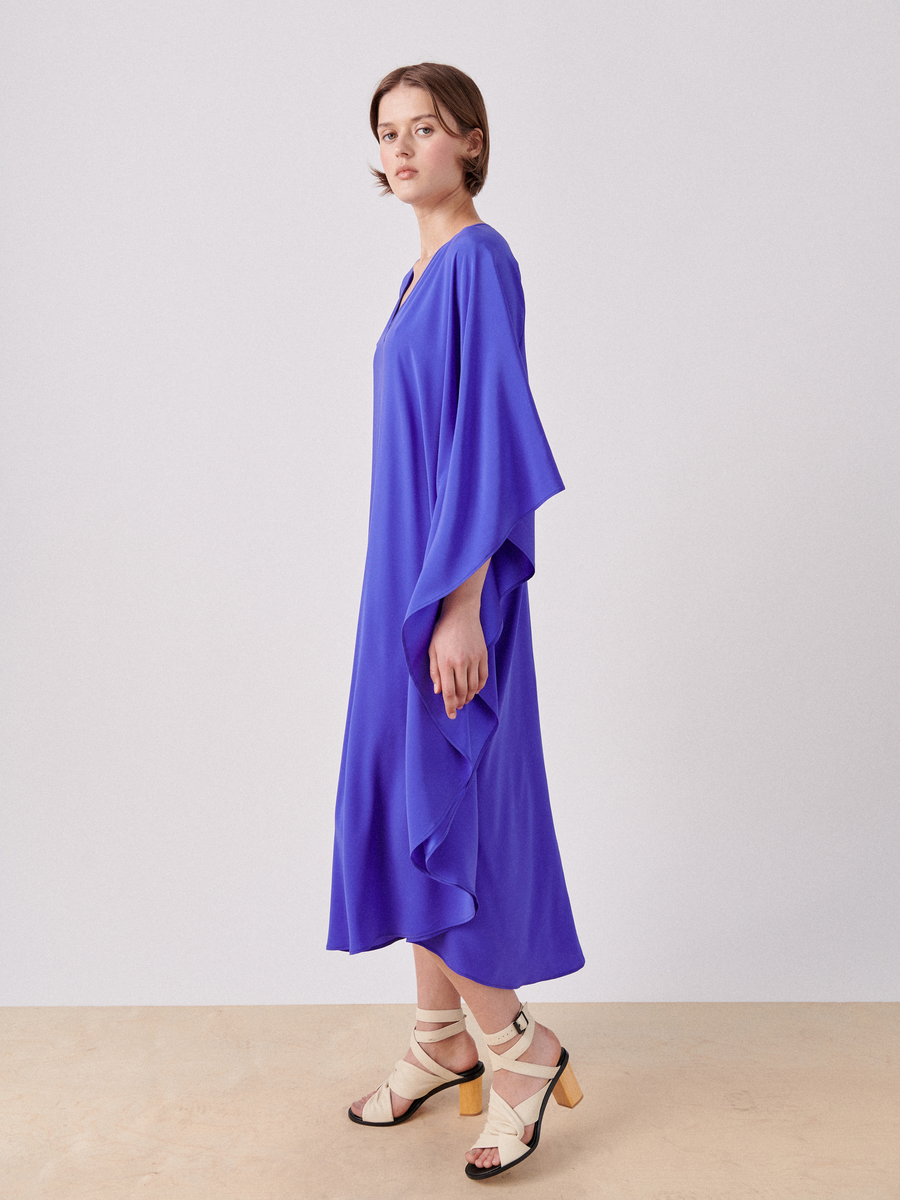 A person stands against a plain background, wearing a loose-fitting, bright blue Circle Caftan by Zero + Maria Cornejo with wide sleeves and beige heeled sandals. They are gazing sideways with a relaxed posture, giving the outfit a shimmering appearance.