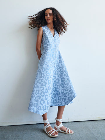 A woman with curly hair wears the Zero + Maria Cornejo Sleeveless Wave Dress—a light blue, animal-print midi dress made from recycled polyester—paired with white strappy sandals, standing against a white wall with hands in her pockets.