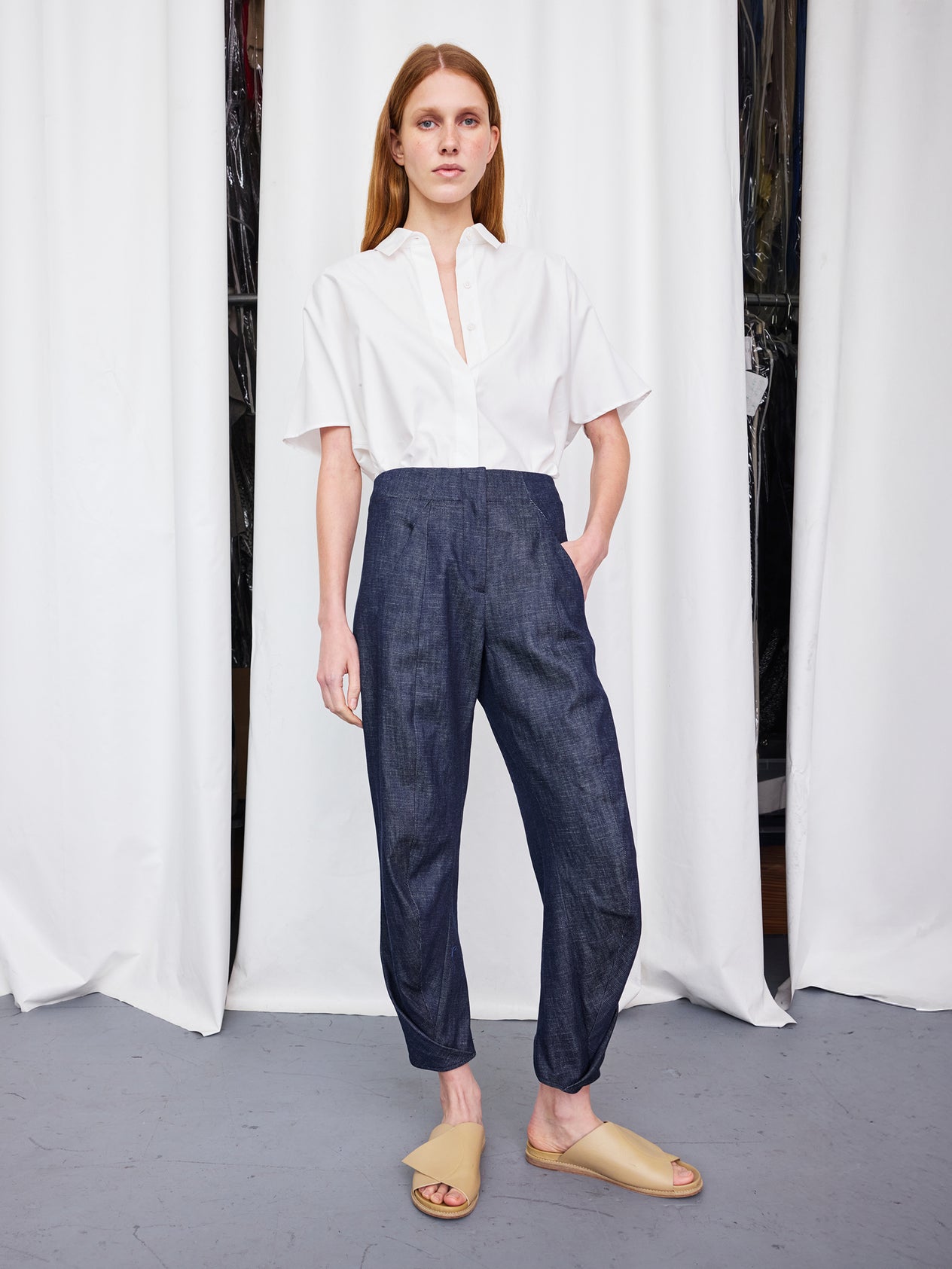 A person with curly hair stands against a white wall, wearing a white blouse and the high-waisted Takeo Pant by Zero + Maria Cornejo, crafted from 100% organic cotton. Their look is completed with white shoes, and they maintain a neutral expression with their hands resting in their pockets.