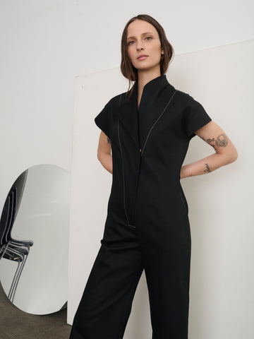 Against a minimal backdrop, a person confidently wears the Edi Boiler Suit by Zero + Maria Cornejo, crafted from GOTS-certified cotton. Tattoos adorn their arms. Beside them, a modern mirror reflects part of a chair, enhancing the polished aesthetic.