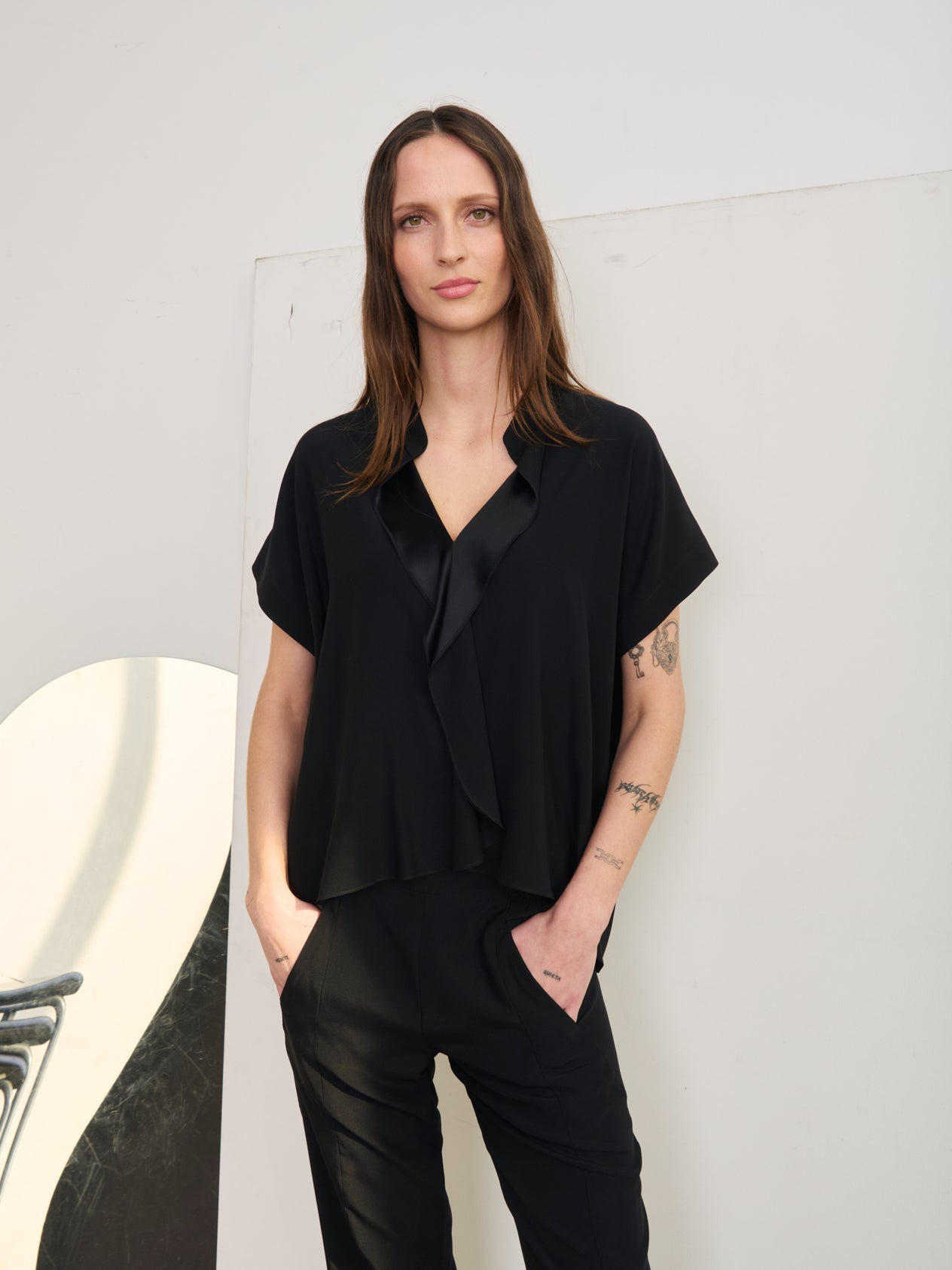 A person with long, curly hair stands against a plain white background, wearing the Fin Gaban Shirt by Zero + Maria Cornejo— a short-sleeved, loose-fitting cream blouse featuring draped ruffle detail— and black pants. A dress form is partially visible on the left side.