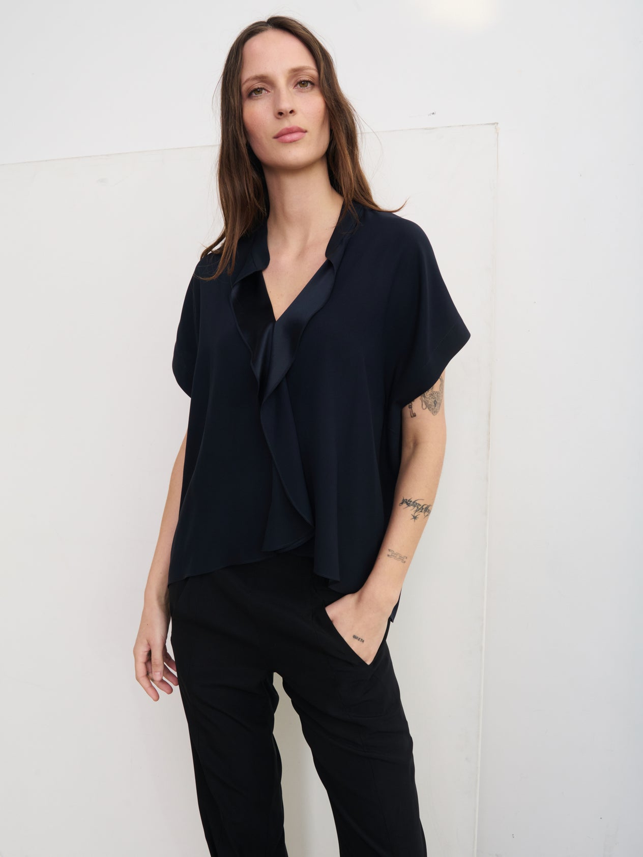 A person with long, curly hair stands against a plain white background, wearing the Fin Gaban Shirt by Zero + Maria Cornejo— a short-sleeved, loose-fitting cream blouse featuring draped ruffle detail— and black pants. A dress form is partially visible on the left side.