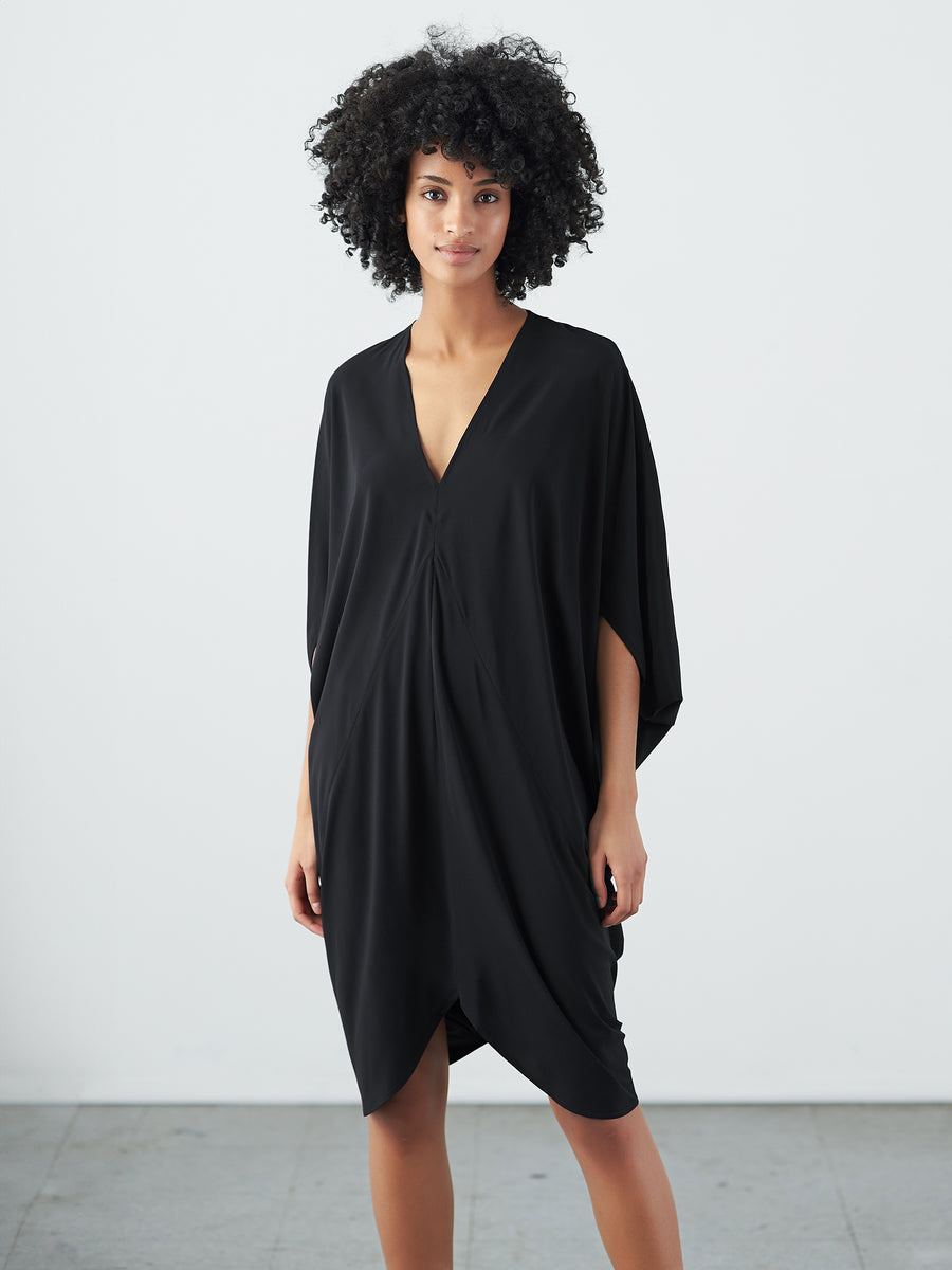 A woman with curly hair wears the loose-fitting Koya Dress in black silk charmeuse, featuring a deep V-neck. She stands on a light gray floor against a plain white background, looking directly ahead. The dress is from the brand Zero + Maria Cornejo.