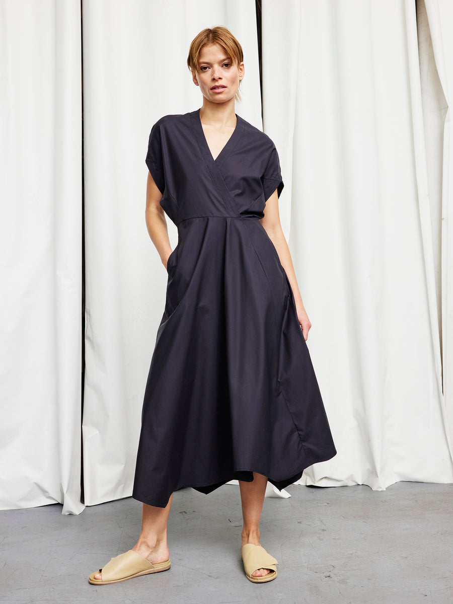 A person stands indoors before white curtains in a relaxed pose, wearing the Zero + Maria Cornejo Long Aki Wave Dress—a loose-fitting, short-sleeved cotton broadcloth style with pockets—paired with beige slip-on shoes.