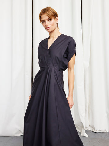 A woman stands before white curtains wearing the Long Aki Wave Dress by Zero + Maria Cornejo, a dark cotton broadcloth piece with a wrap-effect V-neck and a long, flowing skirt. She gazes slightly sideways with a neutral expression.