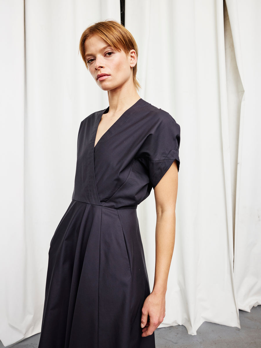 A person with short hair wears the Zero + Maria Cornejo Long Aki Wave Dress, featuring a wrap-effect V-neck, and stands confidently in front of white curtains, looking directly at the camera.