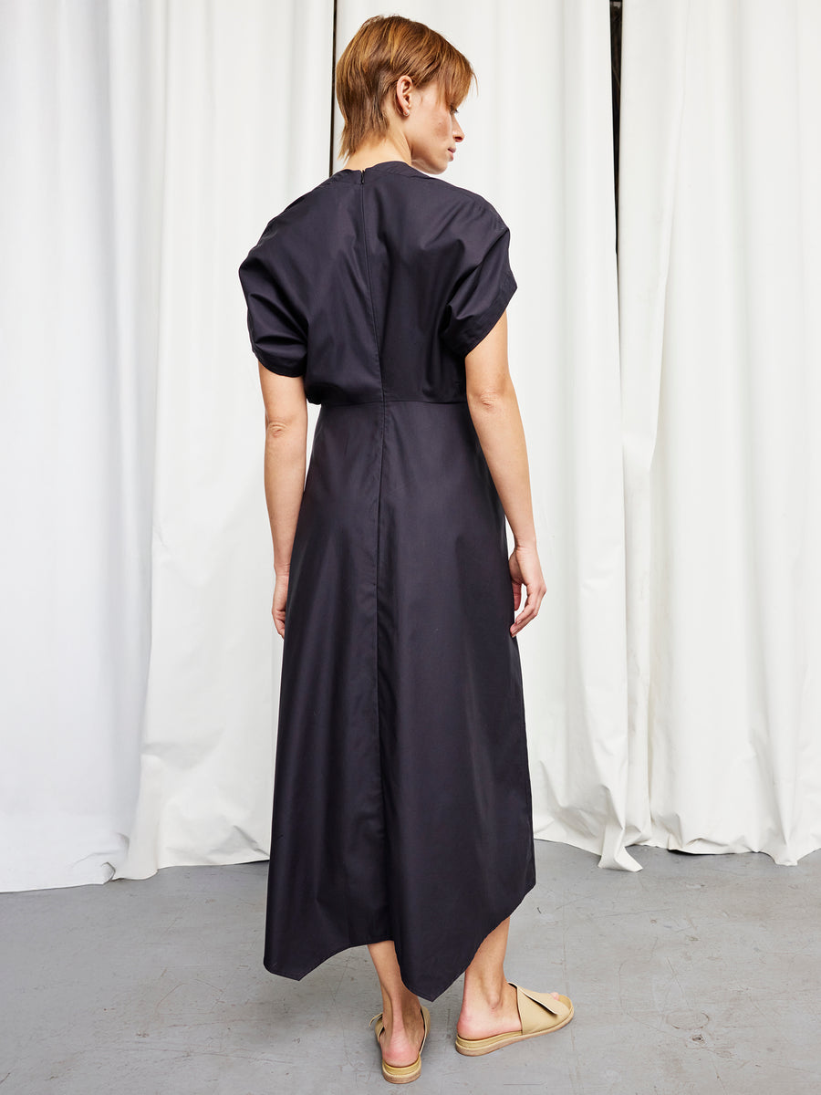 A person stands indoors with their back to the camera, wearing the Zero + Maria Cornejo Long Aki Wave Dress in black cotton broadcloth and beige sandals. White curtains and a gray floor are in the background.
