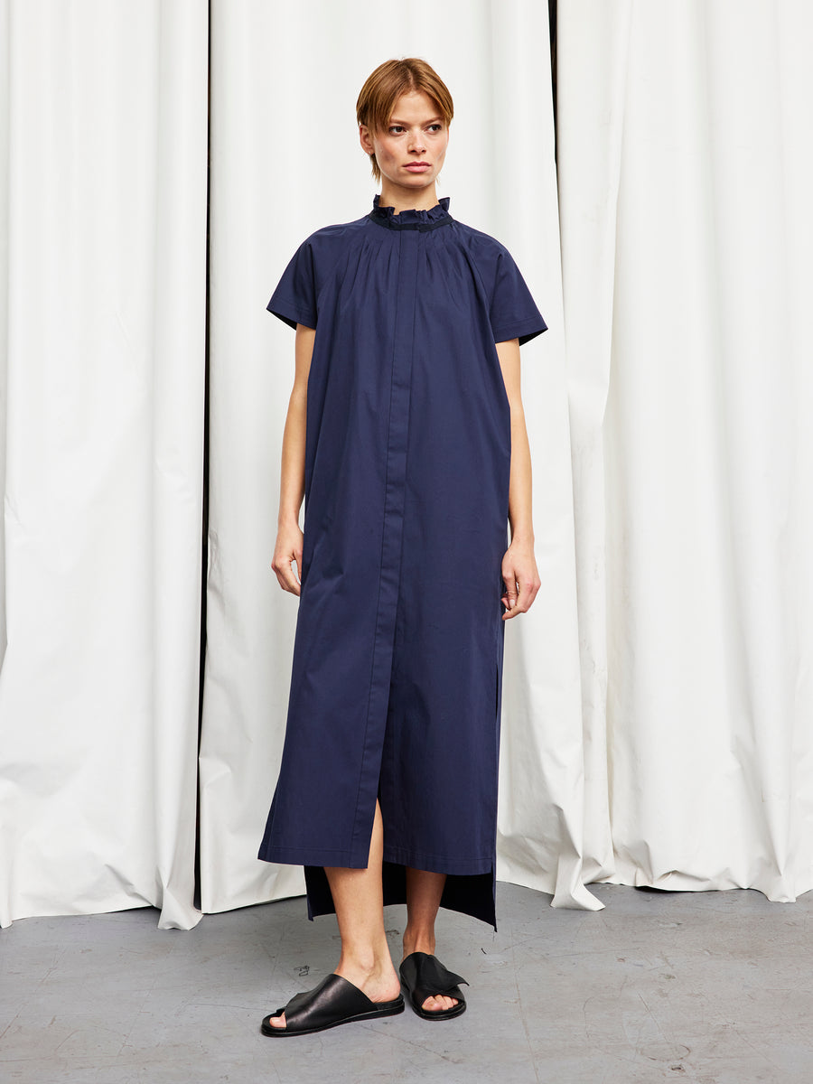 A person stands indoors in a navy Long Pintuck Gaban Dress by Zero + Maria Cornejo, paired with black slide sandals and short hair, set against white curtains.