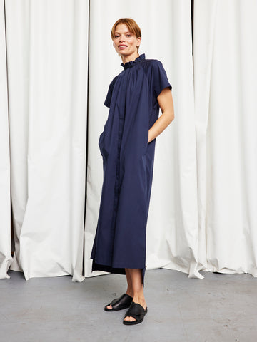 A woman stands before white curtains in the Zero + Maria Cornejo Long Pintuck Gaban Dress—an ankle-length, short-sleeve navy cotton broadcloth style with a ruffled collar and pockets. She pairs it with black sandals. Made in New York.