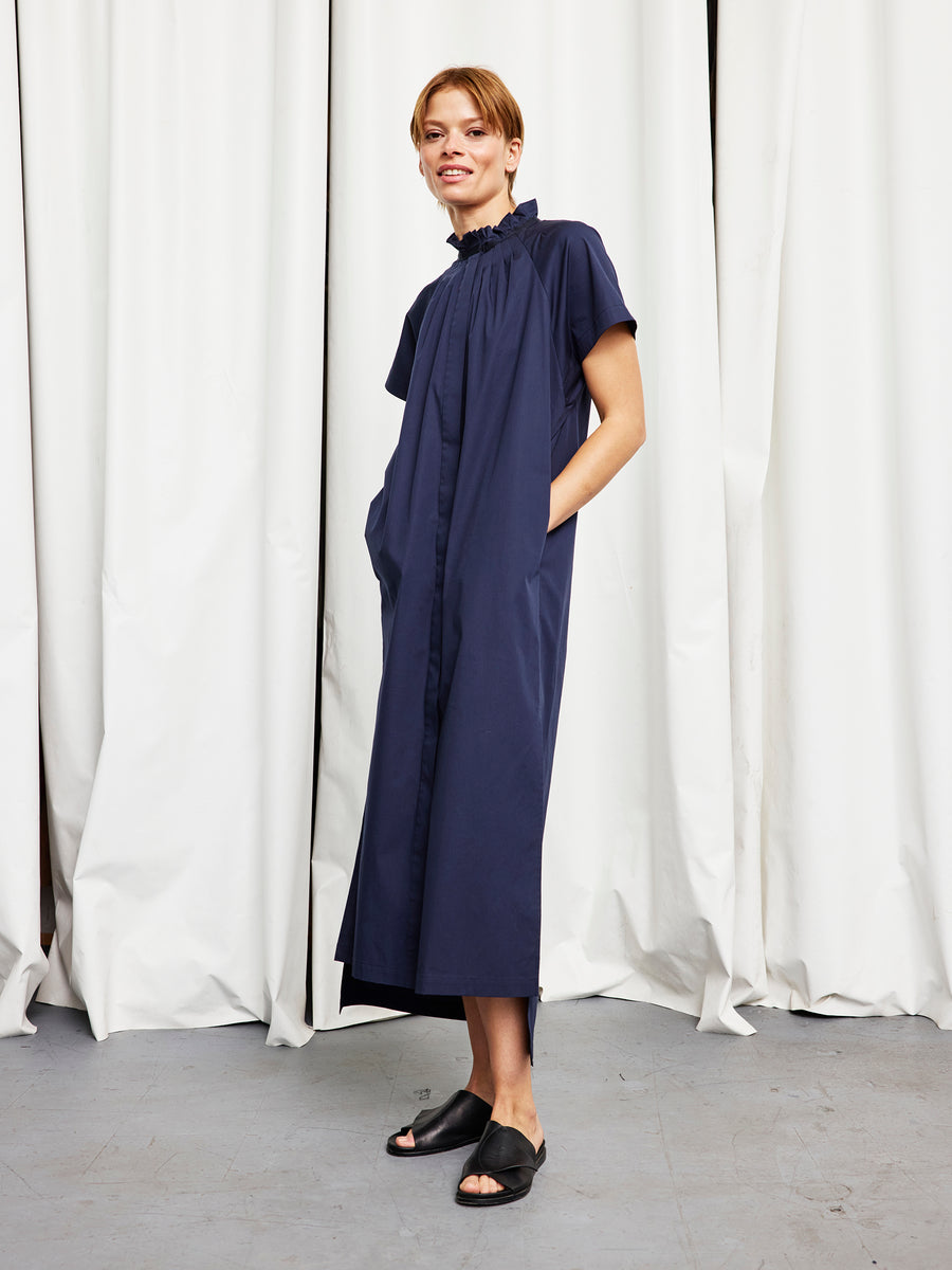 A woman stands before white curtains in the Zero + Maria Cornejo Long Pintuck Gaban Dress—an ankle-length, short-sleeve navy cotton broadcloth style with a ruffled collar and pockets. She pairs it with black sandals. Made in New York.