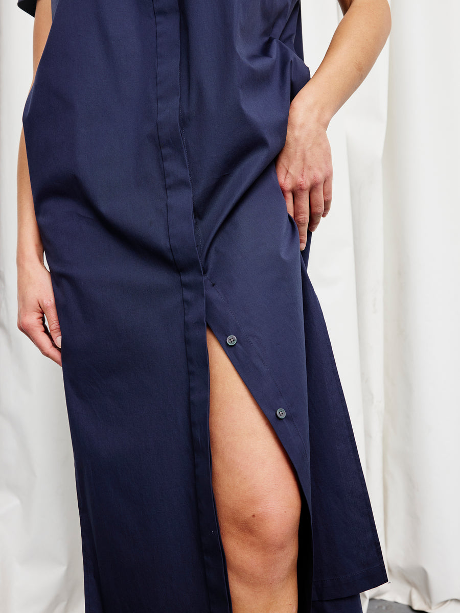 A person stands against a white background wearing the Zero + Maria Cornejo Long Pintuck Gaban Dress in navy blue, mid-length with a thigh-high slit. Their hand rests on the cotton broadcloth, which is made in New York, near the slit.