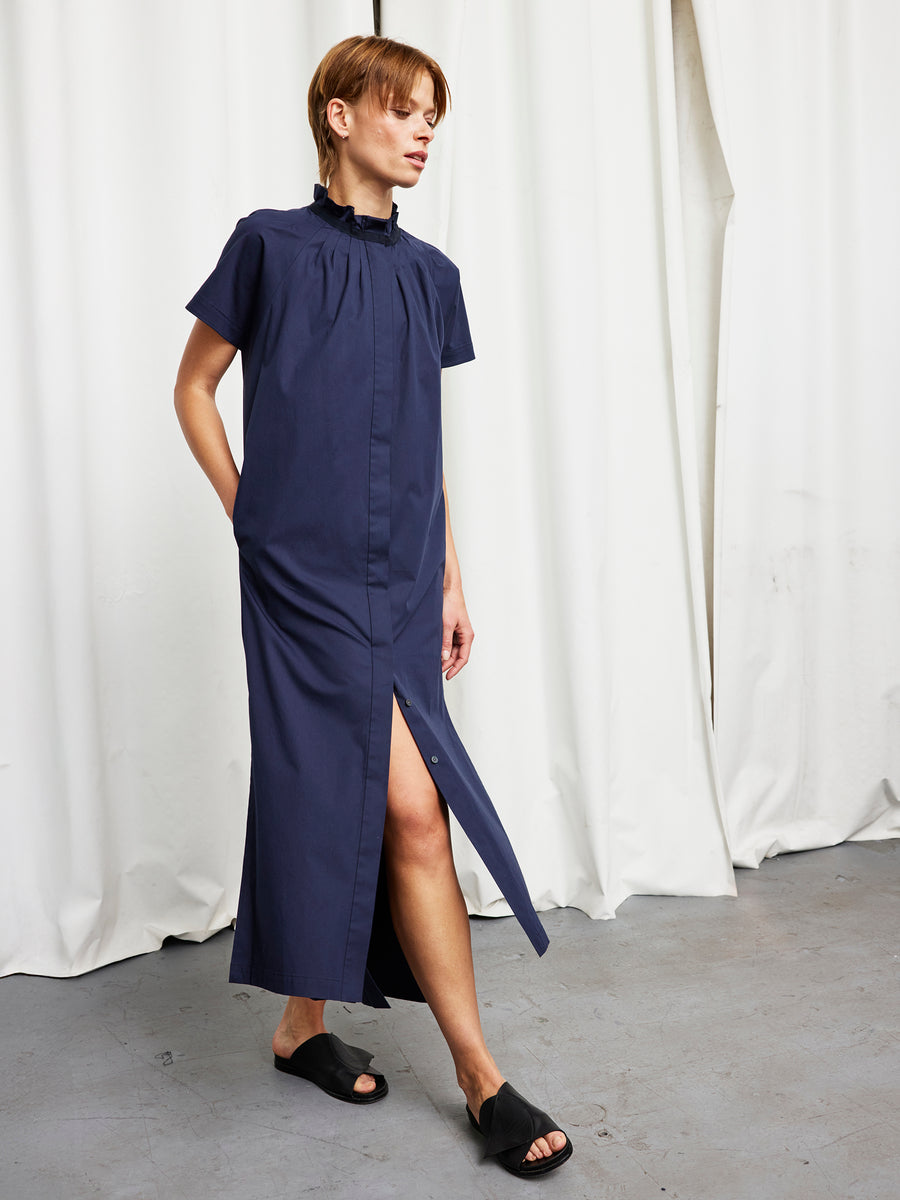 A person wearing the Zero + Maria Cornejo Long Pintuck Gaban Dress and black slide sandals stands in front of white curtains on a gray floor.