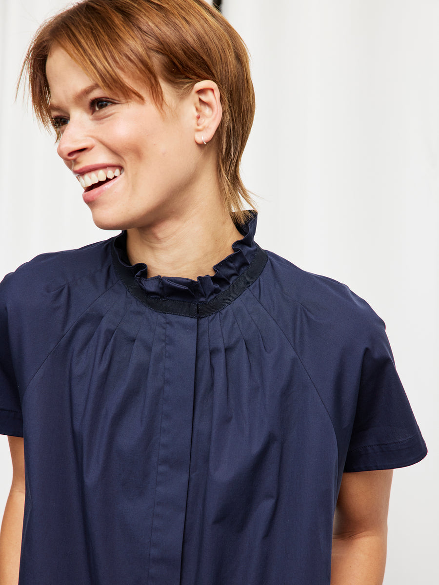 A woman with short brown hair smiles and looks to the side while wearing the Zero + Maria Cornejo Long Pintuck Gaban Dress. The background is softly blurred and white.