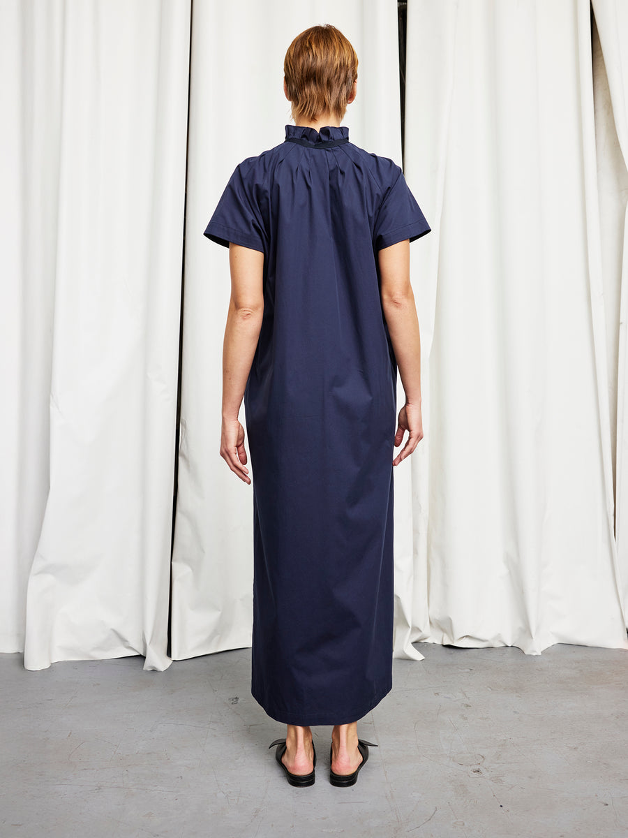 A person with short hair faces away, wearing the Zero + Maria Cornejo Long Pintuck Gaban Dress—a loose navy cotton broadcloth dress with short sleeves and a ruffled collar. White curtains and a gray floor are in the background. Made in New York.