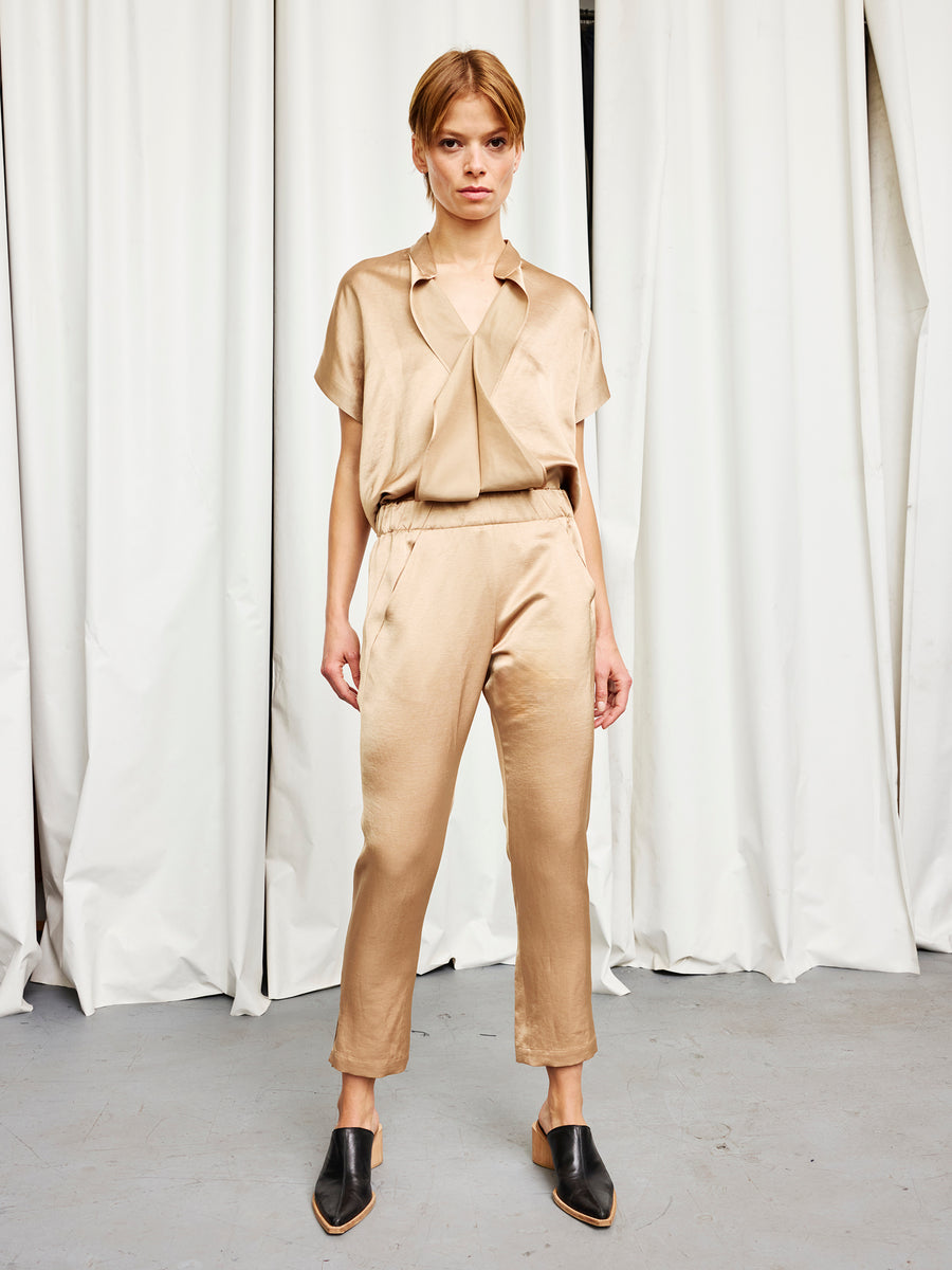 A person stands confidently on a gray floor before white curtains, wearing Zero + Maria Cornejo's Slim Elli Pant in beige with a matching tie-neck blouse and black mule shoes.
