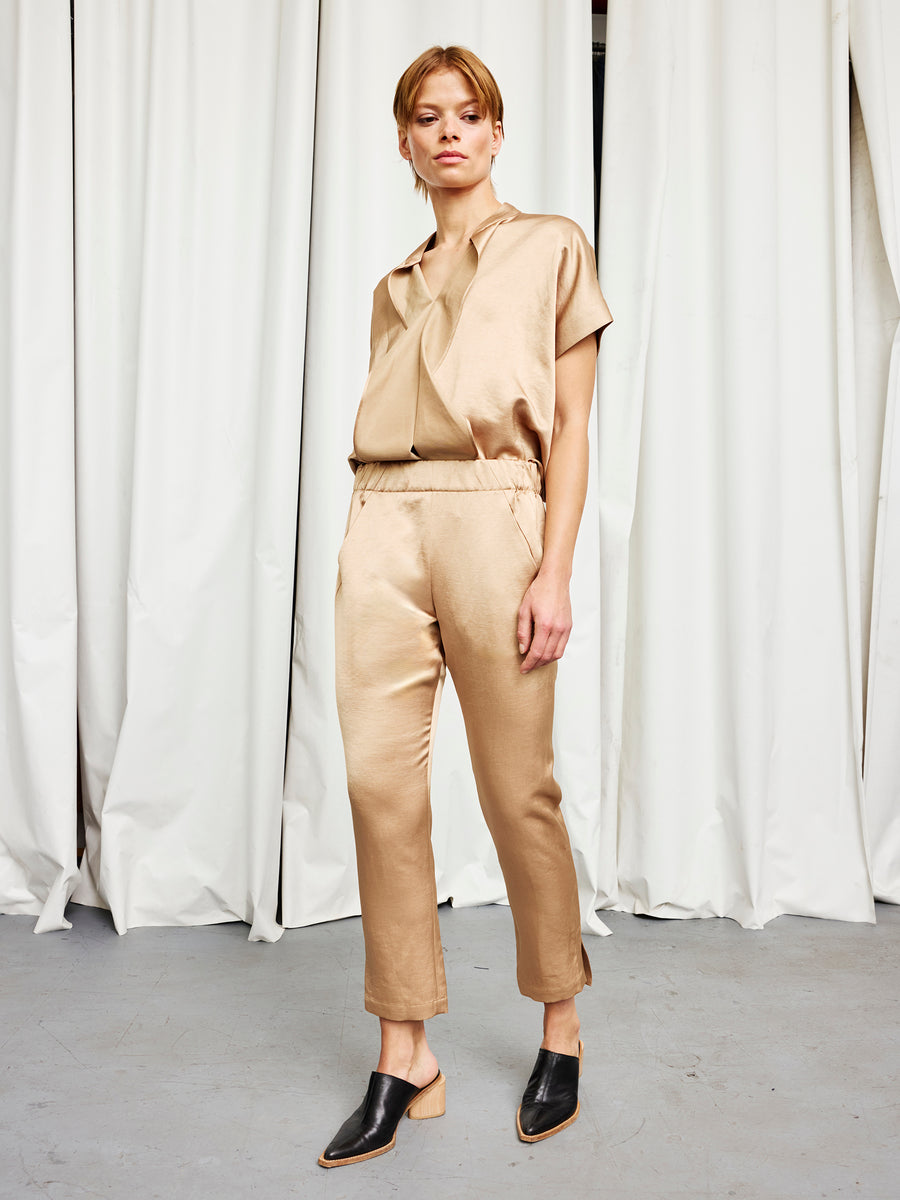 A person stands indoors wearing the Zero + Maria Cornejo Slim Elli Pant and a shiny light brown satin blouse with black slip-on shoes. White curtains and a gray floor are in the background.