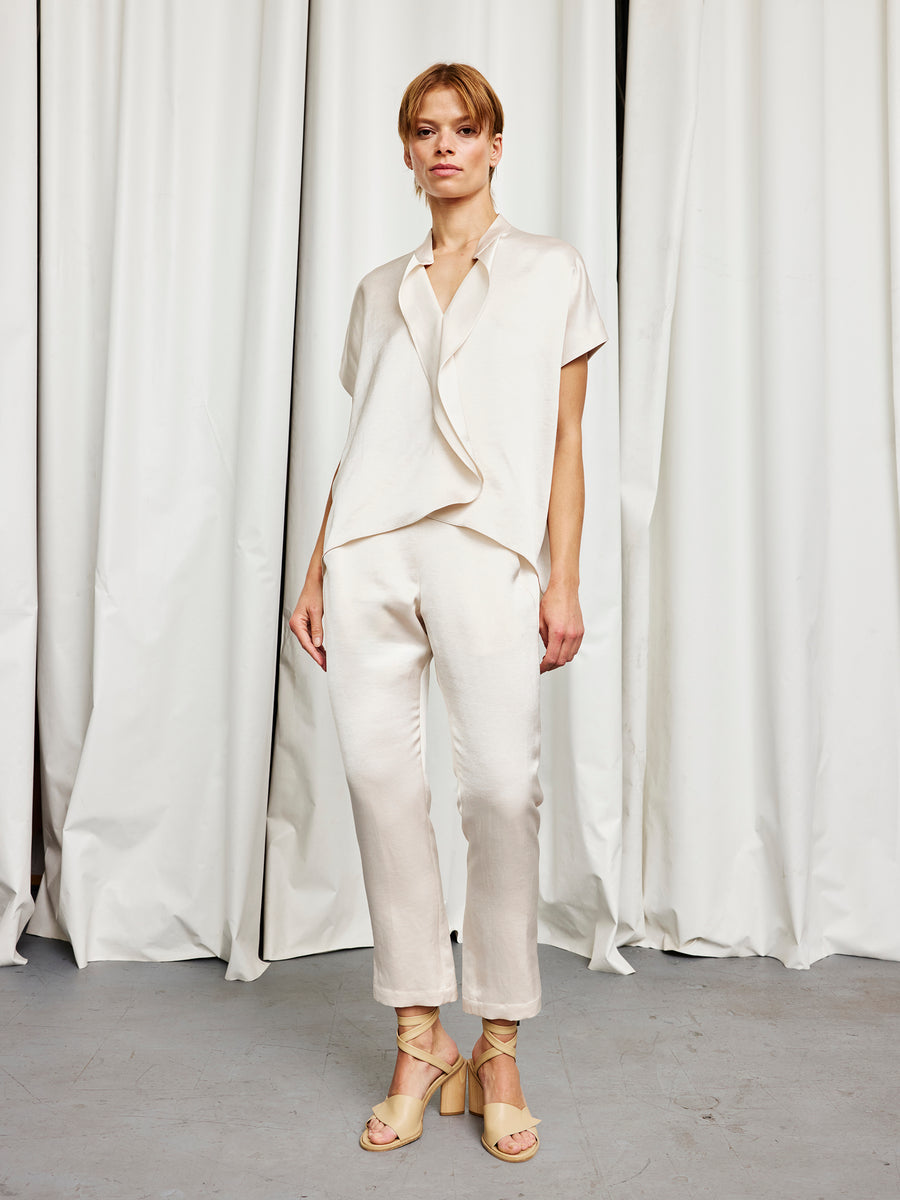 A person with short hair stands before white curtains in a loose cream short-sleeve blouse, beige strappy sandals, and Zero + Maria Cornejo Slim Elli Pant featuring a straight, relaxed leg.