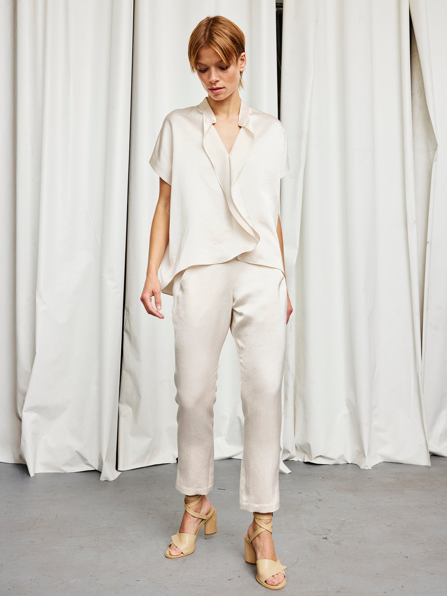 A person stands indoors before white curtains, wearing a loose light beige blouse, the Slim Elli Pant by Zero + Maria Cornejo, and beige open-toe heeled sandals. They look down with hands at their sides.