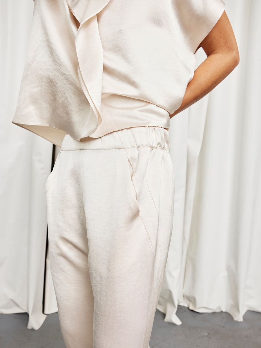 A person poses with one hand on their hip, wearing Zero + Maria Cornejo's Slim Elli Pant—a pair of cream-colored, relaxed-fit linen-blend trousers—paired with a loose blouse. White flowing curtains hang in the background.