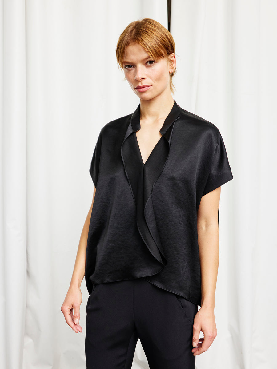 A person with short, light brown hair stands before white curtains, wearing the Zero + Maria Cornejo Fin Gaban Shirt in black and black pants.