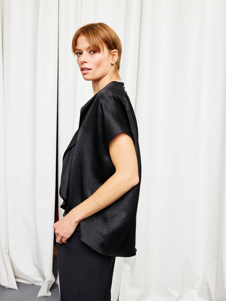 A person with short light brown hair stands sideways in front of white curtains, wearing the Black Fin Gaban Shirt by Zero + Maria Cornejo and loose black pants.