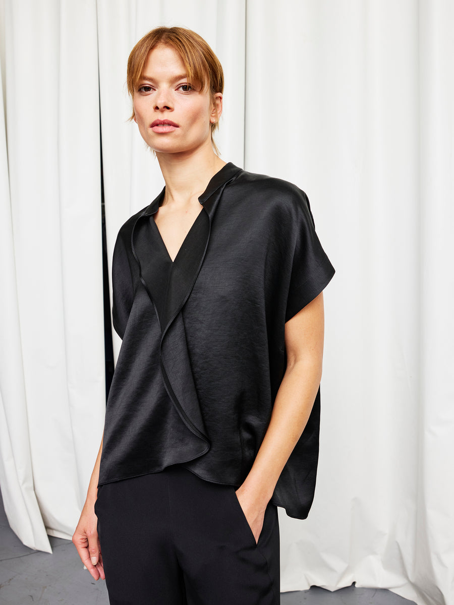 A person stands before white curtains wearing the Fin Gaban Shirt by Zero + Maria Cornejo—a loose, short-sleeved, black linen-blend shirt layered over a black top and pants, with one hand in their pocket.