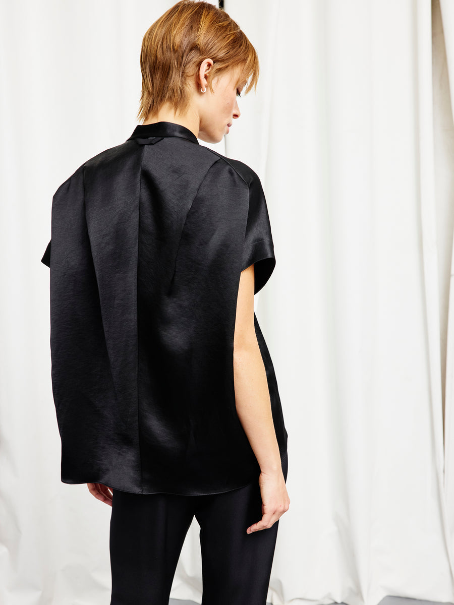 A person with short light brown hair, wearing the Zero + Maria Cornejo Fin Gaban Shirt in black and matching pants, stands facing away before white curtains.