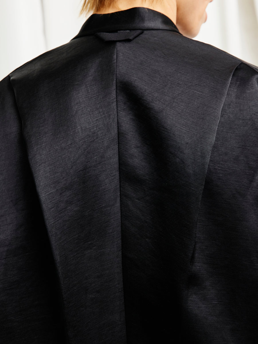 Close-up of the back of a person wearing the Zero + Maria Cornejo Fin Gaban Shirt, showcasing its black, smooth fabric and center seam. A softly blurred background highlights the stand collar and partially visible head and neck.